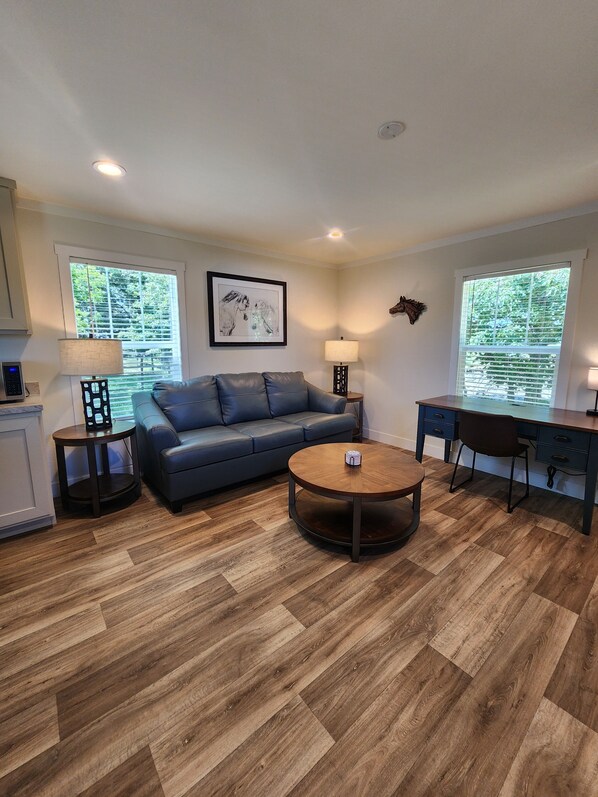 A modern living room with a blue sofa, a round coffee table, and a desk with a chair. There are two windows with blinds, a framed artwork on the wall, and a small table with a lamp.