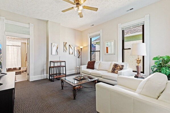 A living room with a white sofa, a glass coffee table, a ceiling fan, and artwork on the walls.