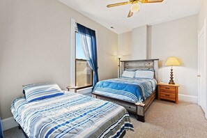 A bedroom with two beds, a ceiling fan, a window with curtains, bedside lamps, and a door.