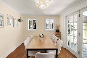 A dining room with a wooden table, white chairs, a chandelier, and large windows.