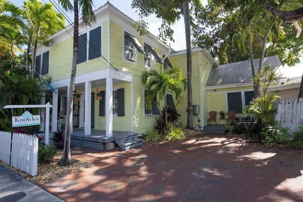 A two-story yellow house with a porch, surrounded by palm trees and a brick pathway.