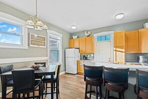 A modern kitchen with wooden cabinets, a white refrigerator, a dining table with chairs, and a hanging light fixture.