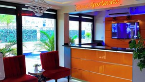 Zona de recepción de hotel con diseño moderno, que incluye un mostrador de recepción, dos sillas rojas, un televisor y vistas al exterior a través de amplias ventanas.