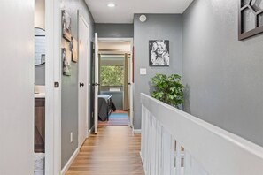 A hallway with a wooden floor, a white railing, and a door leading to a room with a bed and a window.