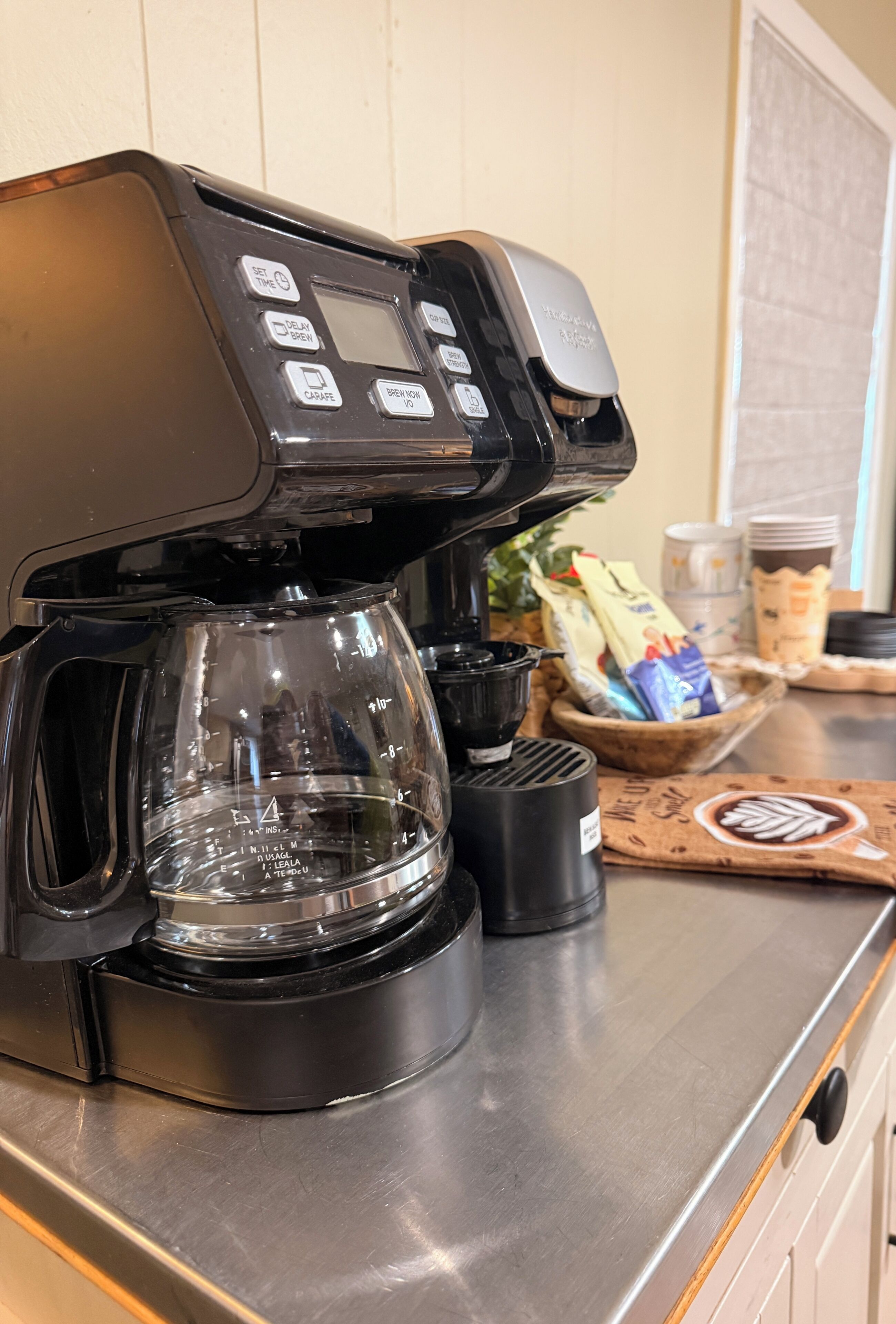A coffee machine with a glass carafe and a drip tray, a bag of coffee pods, and a stack of plates on a counter.