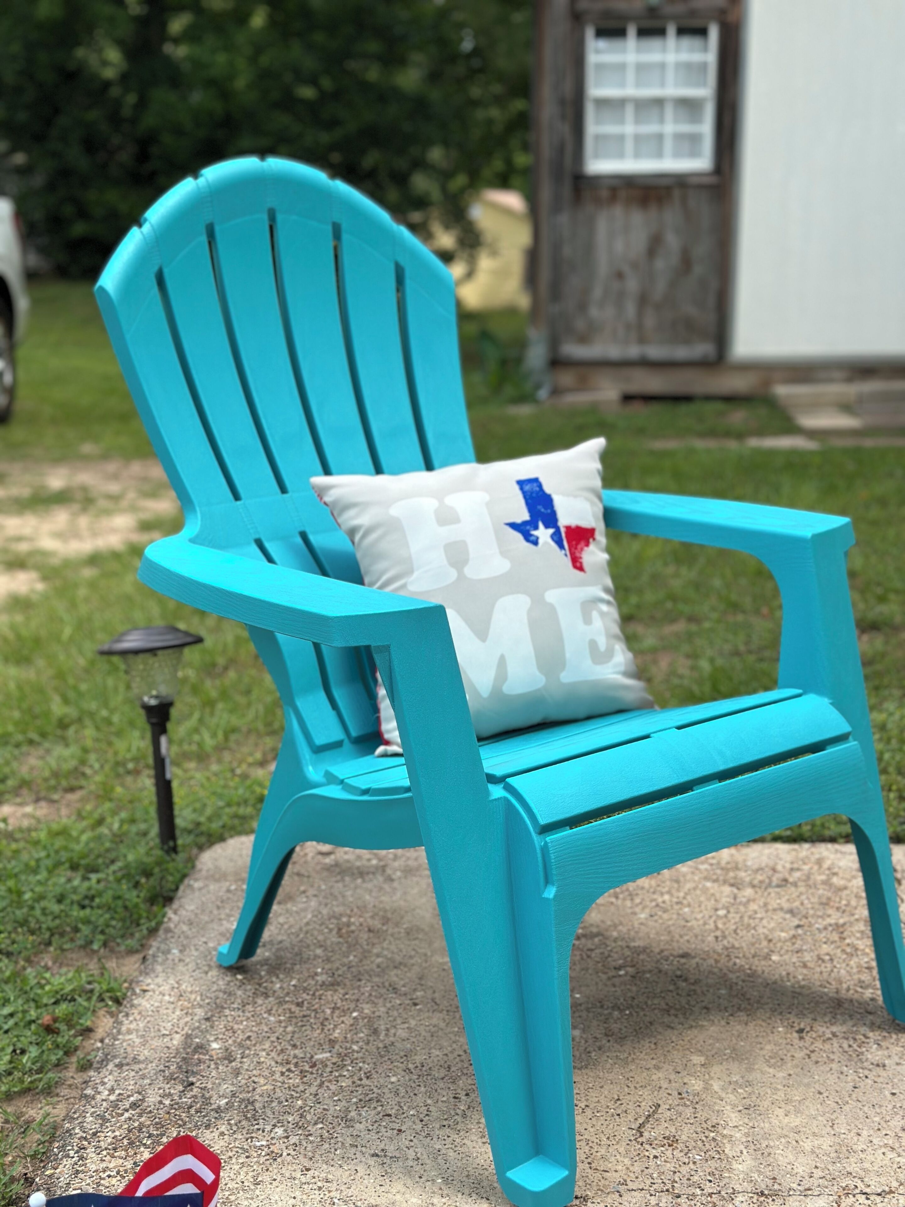 Turkoosi Adirondack-tyylinen puutarhatuoli, jossa on koristetyyny, jonka kuvioissa on sana »KOTI» ja Texasin osavaltion lippu, on sijoitettu betoniterassille.