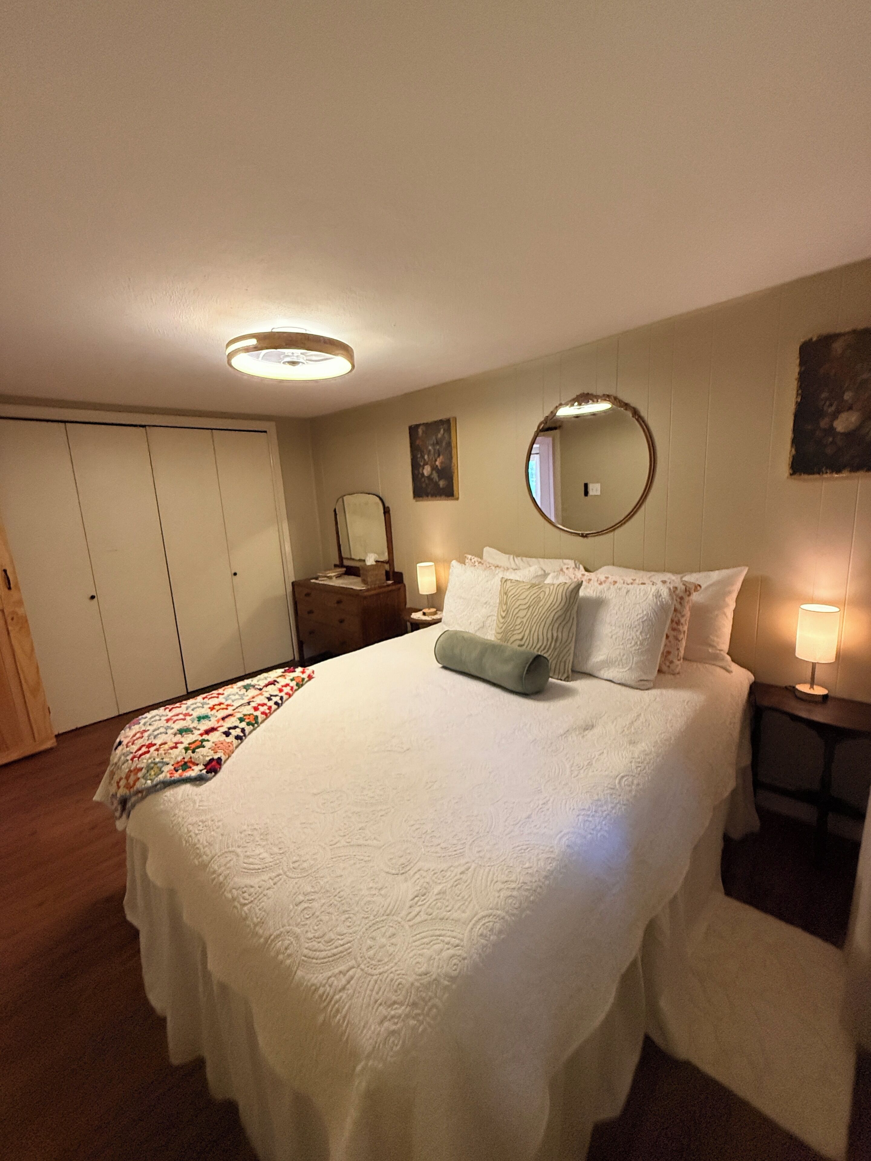 Ein Hotelzimmer mit einem großen Bett, einem Nachttisch mit Lampe, einem runden Spiegel, einem Kleiderschrank und einer blumengemusterten Tagesdecke auf dem Bett.