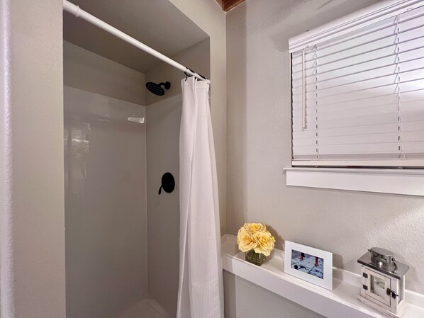 A bathroom with a shower, a white towel, a black shower head, a white window with blinds, a vase with yellow flowers, and a framed picture on a shelf. - Country Chic 1 BR apartment on 2 acres w/charming historic patio in serene Azle (Azle)