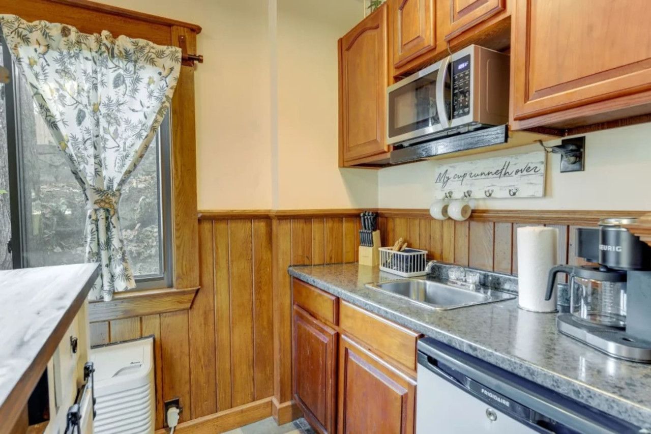 A compact kitchen with wooden cabinets, a microwave, a coffee maker, and a window with a floral curtain.