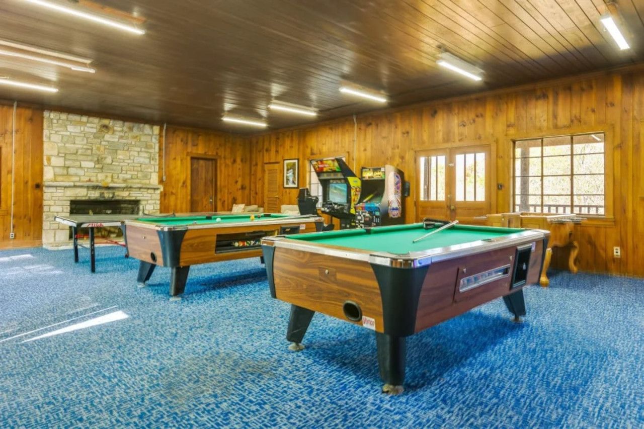 A room with three pool tables, a stone fireplace, wooden walls, and large windows.