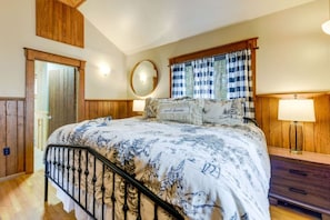 A bedroom with a bed, wooden walls, a window with checkered curtains, a bedside table with a lamp, and a mirror on the wall. - Village 37 Stone Cabin with Private Hot Tub (Bryson City)