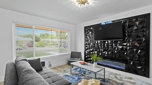 A modern living room with a grey sectional sofa, a flat-screen TV mounted on a geometric wall, and a coffee table with a plant. - Incredible 4-bedroom home in Tequesta FL with AC, WiFi and absolute comfort. (Tequesta)