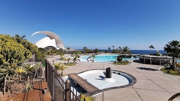 Zwembadgedeelte van een resort met een grote, gebogen witte constructie op de achtergrond, omringd door palmbomen en een helderblauwe lucht.
