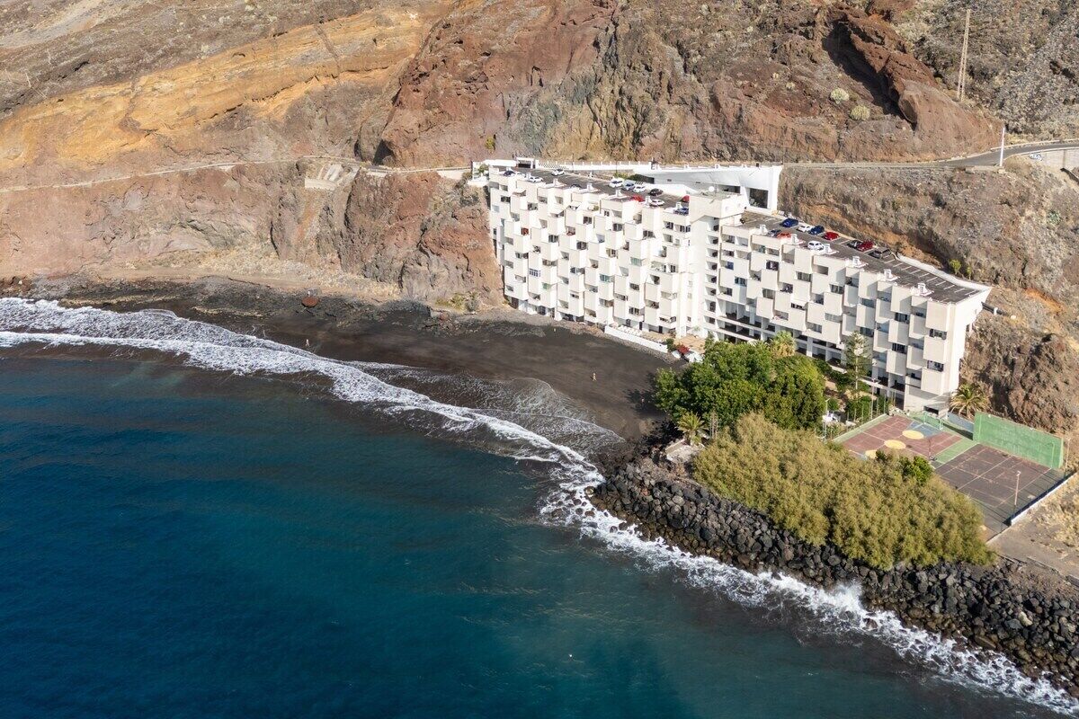 Vue aérienne d’un hôtel situé sur la côte, avec un rivage rocheux, un court de tennis et un parking.