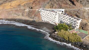 Vue aérienne d’un hôtel situé sur la côte, avec un rivage rocheux, un court de tennis et un parking.