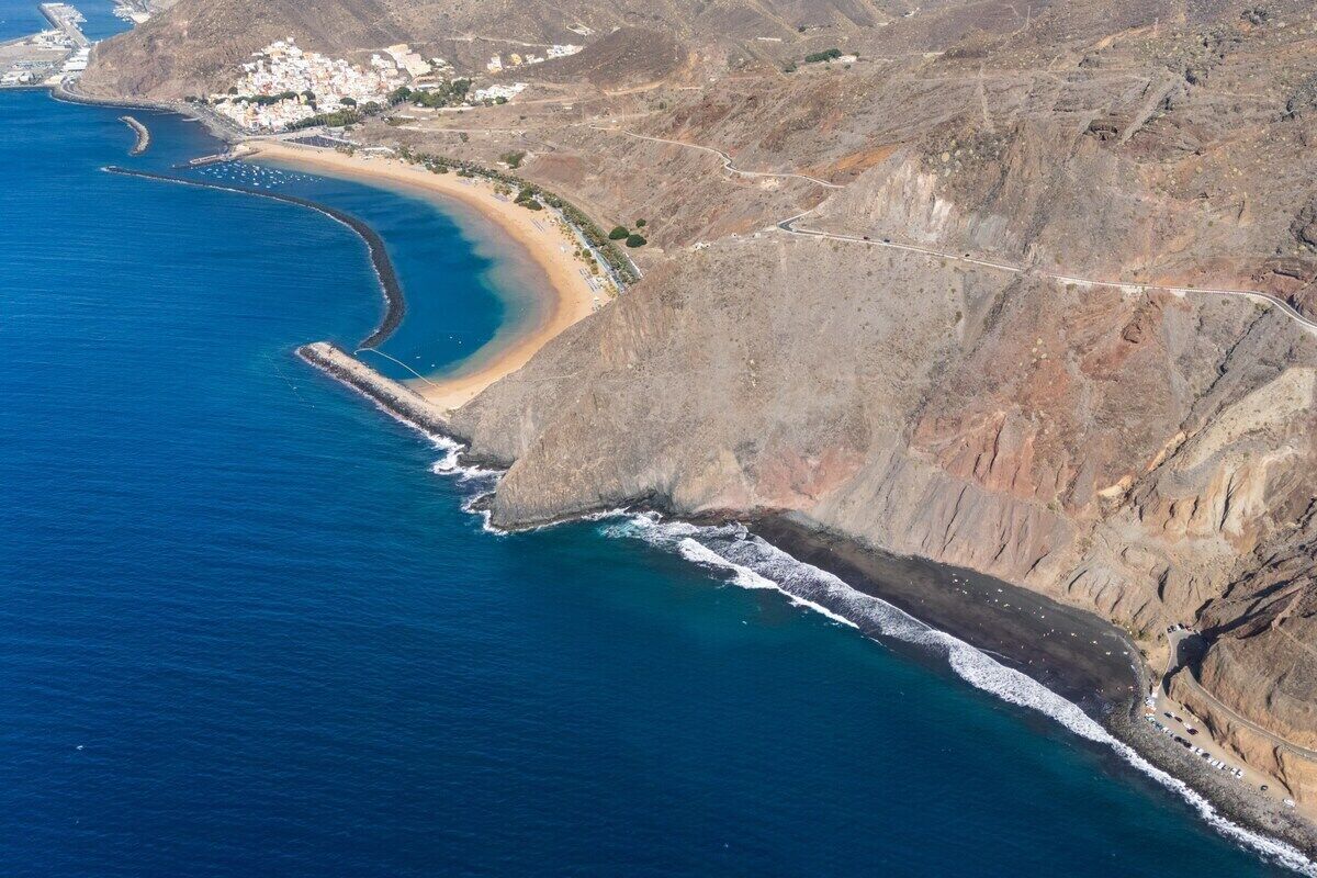 An aerial view of a coastal area with a beach, rocky cliffs, and a road curving along the coastline.