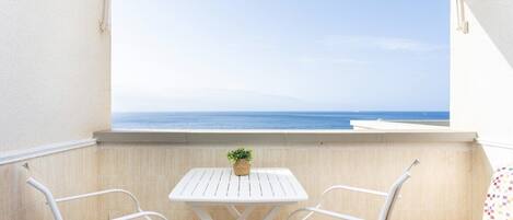A balcony with a white table and two chairs, a small plant, and a view of the sea.