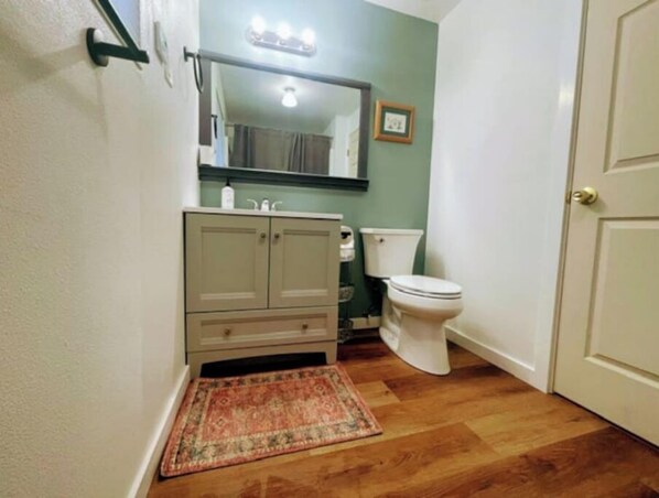 Une salle de bain avec un plancher en bois, des toilettes blanches, un meuble beige et un petit tapis.