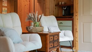 Une pièce chaleureuse avec deux fauteuils blancs, une table d’appoint en bois surmontée d’un panier en osier, et un buffet vert contenant de l’ustensile de cuisine.