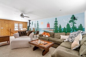 Ein gemütliches Wohnzimmer mit Kamin, Holzbalkendecke und einem Wandbild von Leuchtturm und Wald.