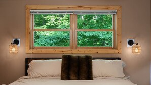 Un lit avec une tête de lit en bois, une fenêtre donnant sur un paysage verdoyant et des appliques murales.
