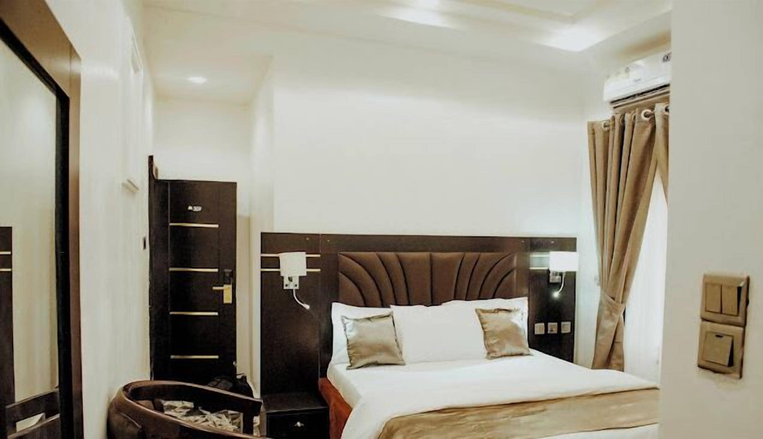 Una habitación de hotel con una cama grande, un armario de madera oscura, una mesita de noche y una ventana con cortinas.