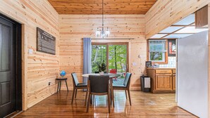 Interior de una cabaña de madera con comedor, cocina y grandes ventanales que ofrecen vistas al exterior.