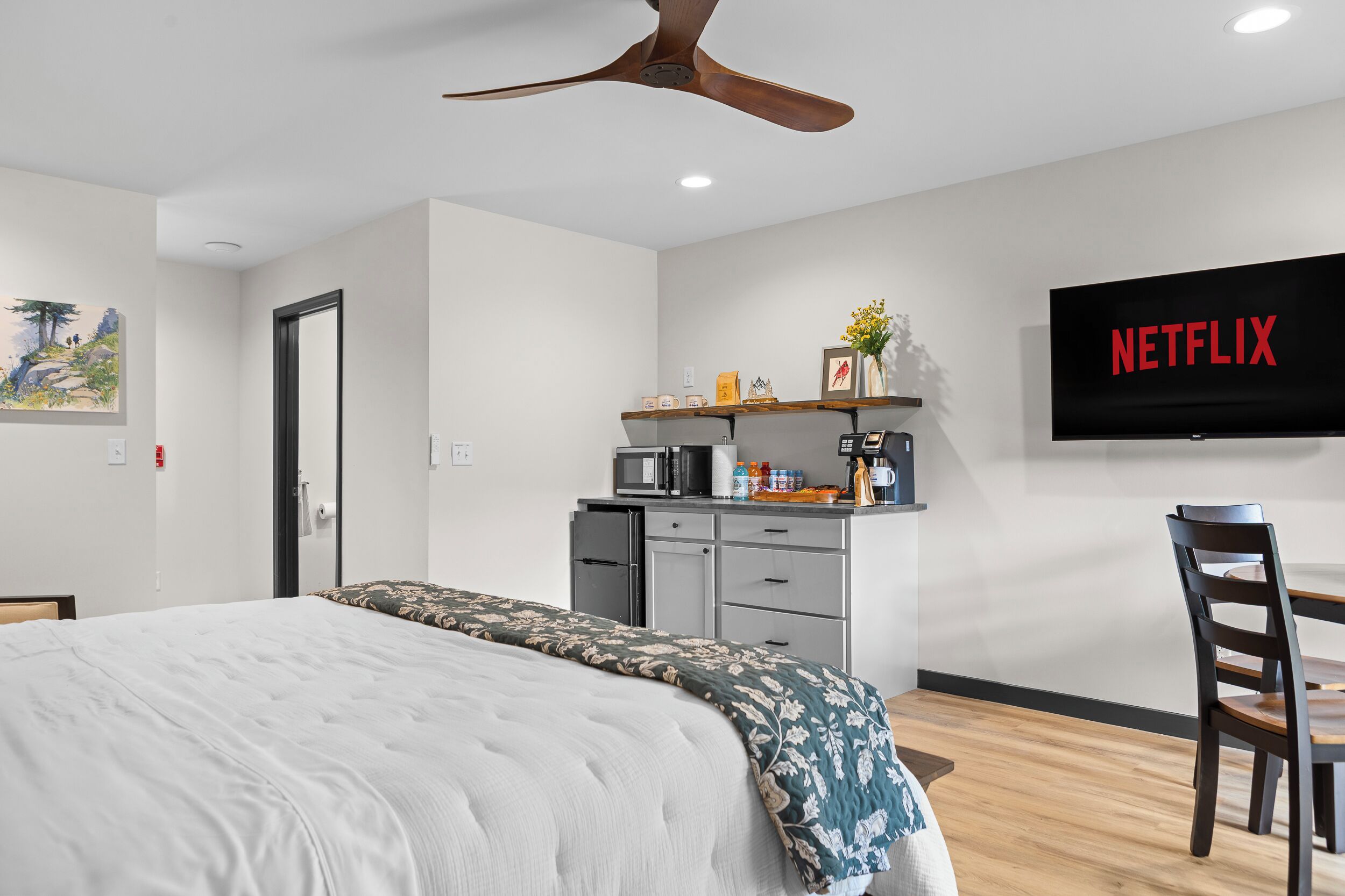 Una habitación de hotel moderna con una cama, un escritorio con silla, ventilador de techo, televisión y una pequeña cocina.