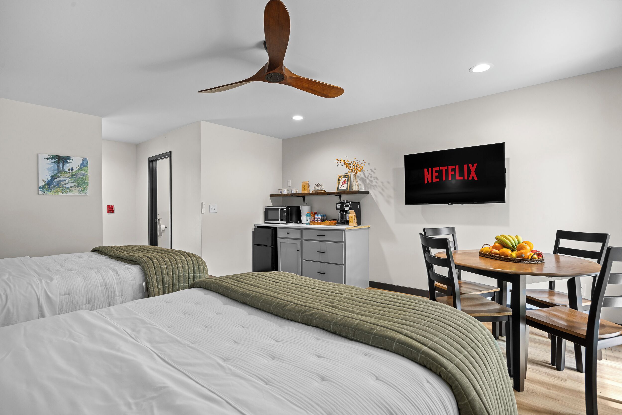 A hotel room with two beds, a ceiling fan, a TV, a desk with a chair, and a dining table with fruit.