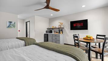 Habitación de hotel con dos camas, ventilador de techo, televisor, escritorio con silla y una mesa de comedor con fruta.