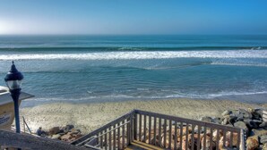 Vista de playa con una escalera de madera que baja a la orilla, una torre de salvavidas y un cielo azul despejado.