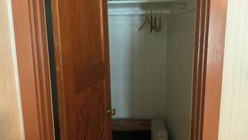 Un pasillo estrecho con una puerta de madera, un estante blanco con objetos colgando y un pequeño cofre de madera.