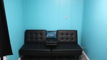 Un canapé noir avec un coussin orné d’un motif de feuilles, adossé à un mur de couleur turquoise, une petite table et une prise électrique sur le mur.