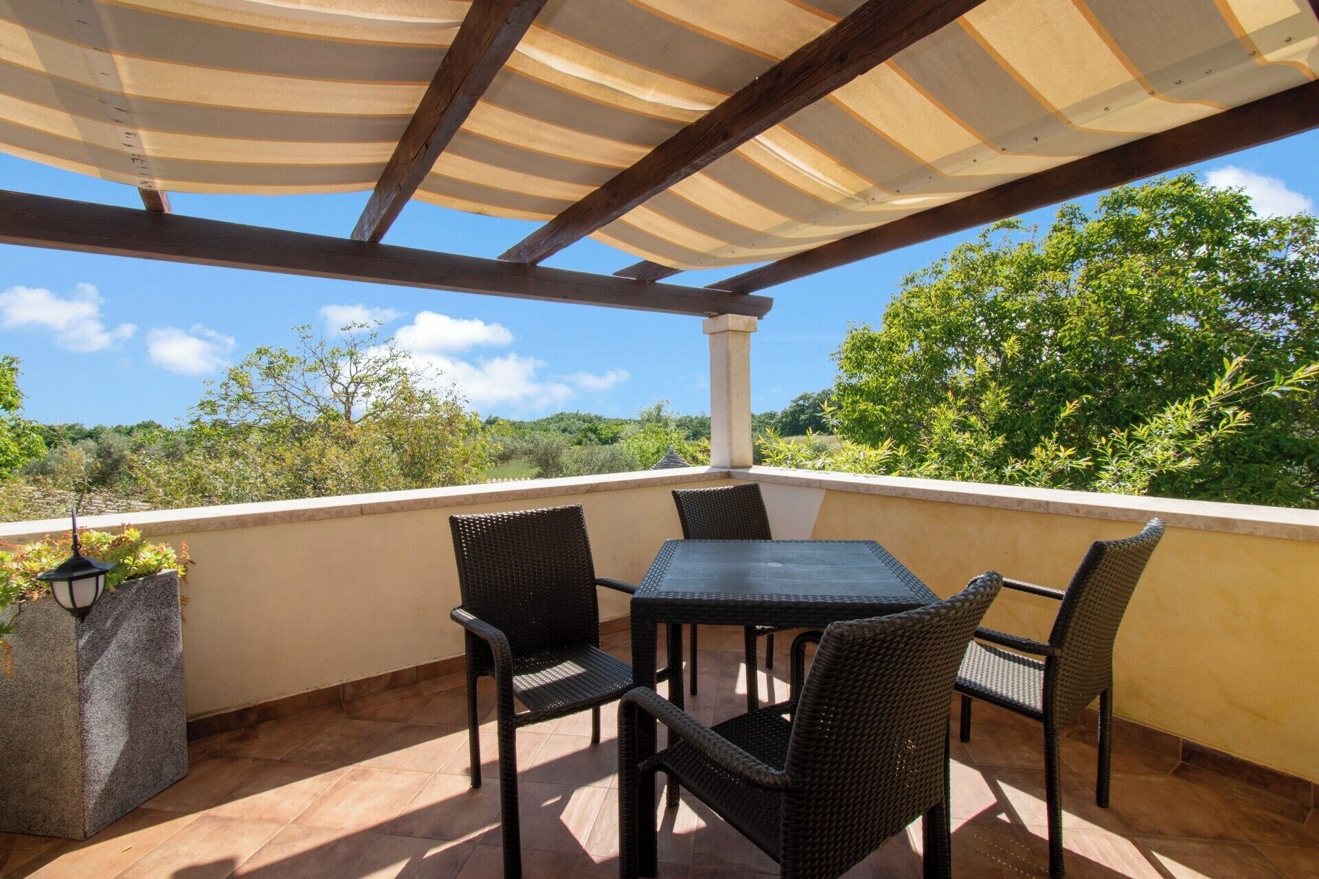 A covered outdoor patio with a table and chairs, a lamp, and a view of greenery and hills.