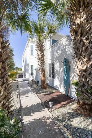 A white building with a blue door, surrounded by palm trees and a gravel pathway.