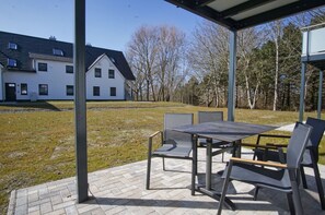 An outdoor patio with a table and chairs, a covered area, and a view of a white building and trees. - Beachfront vacation rental with a terrace, access to the pool and sauna - Rex Rugia - Rex (Göhren)