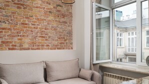 Un salon moderne avec un mur en briques, un canapé beige, une petite table ronde et une fenêtre donnant sur des immeubles.