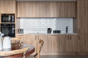A modern kitchen with wooden cabinets, a white tiled backsplash, a built-in oven, a coffee maker, and a dining area with a wooden table and chairs. - The Octagon House (Dunedin)