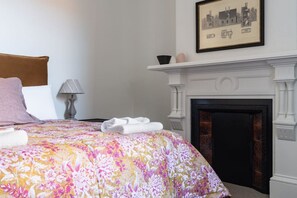 A hotel room with a bed, a fireplace, a framed picture on the wall, and a nightstand with a lamp. - The Octagon House (Dunedin)