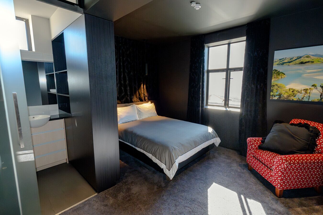 A bedroom with a bed, a red patterned chair, a window with a view, and a built-in wardrobe.