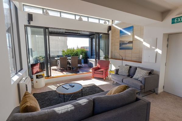 Un salon moderne avec un canapé gris, une petite table ronde et un fauteuil rouge à motifs. Une grande fenêtre laisse entrer la lumière naturelle et offre une vue sur l’extérieur.