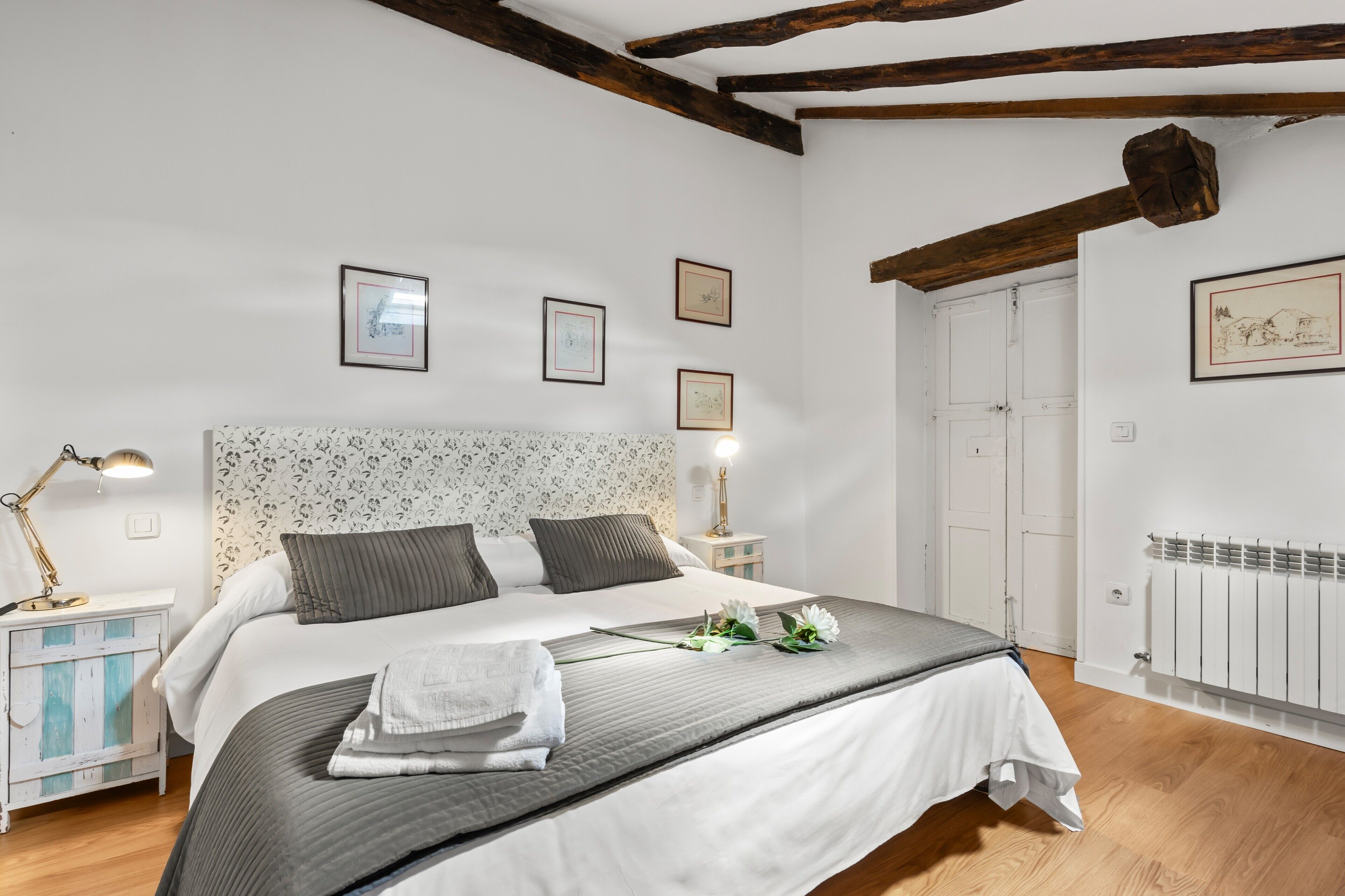 A bedroom with a large bed, white walls, wooden beams, framed pictures, and a radiator.