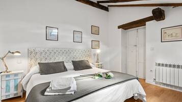 A bedroom with a large bed, white walls, wooden beams, framed pictures, and a radiator.
