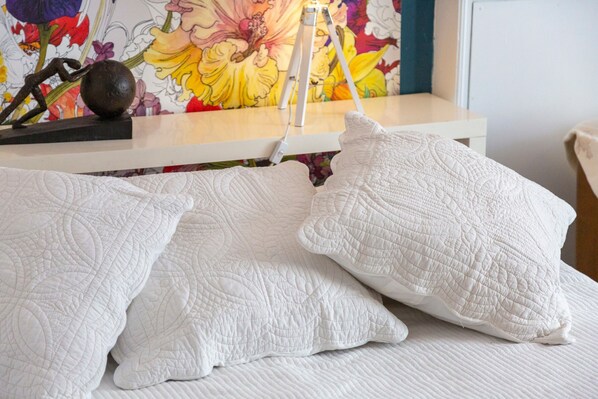 Un lit avec une couette et des draps blancs matelassés, une lampe blanche et, en arrière-plan, un tableau floral aux couleurs vives.