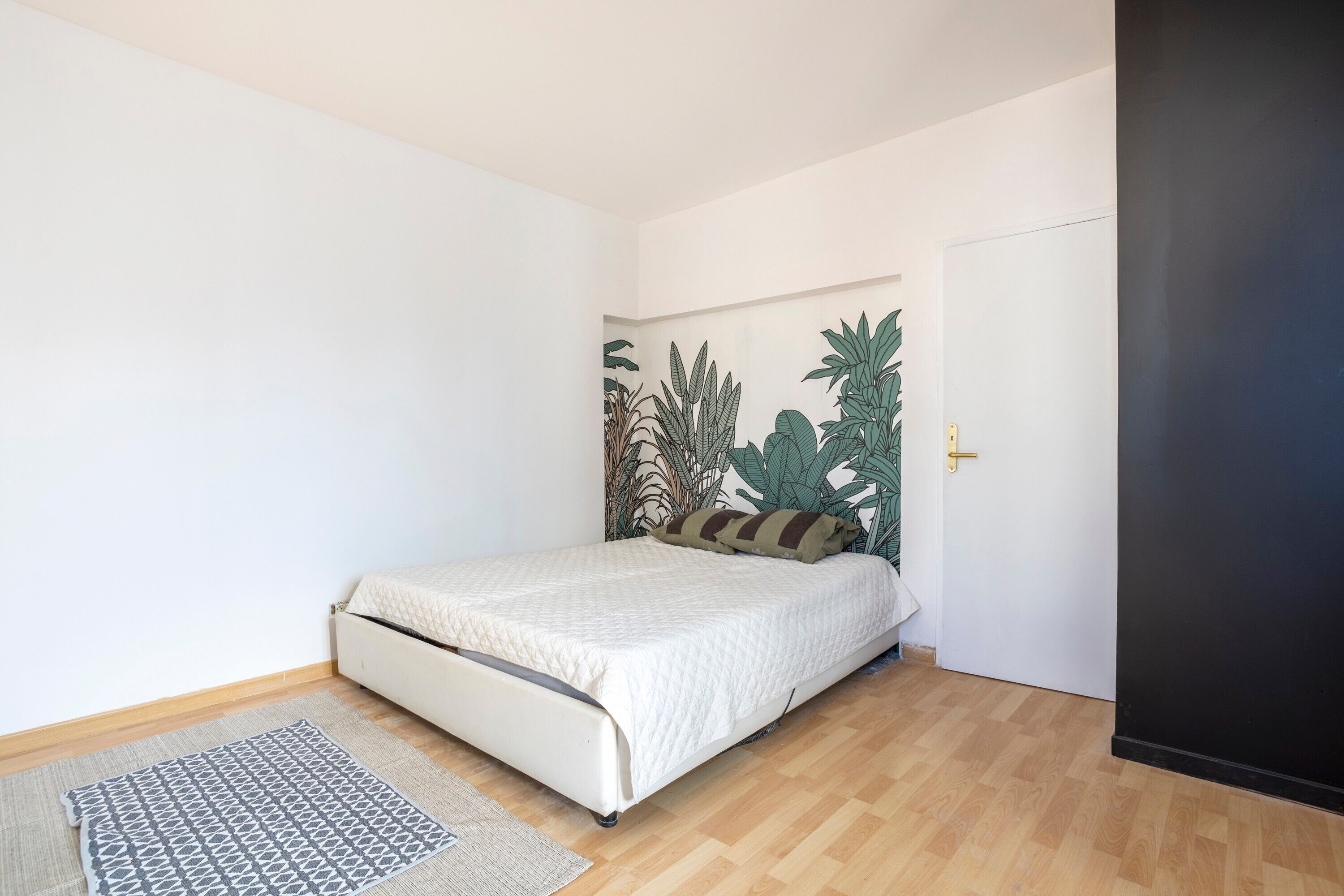 A bedroom with a bed, a patterned rug, a door, and a wall with a plant mural.