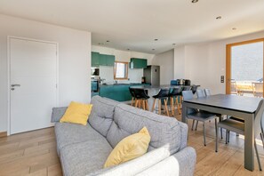 Un salon moderne avec un canapé gris, des coussins jaunes, une table à manger avec des chaises, et une cuisine en arrière-plan.