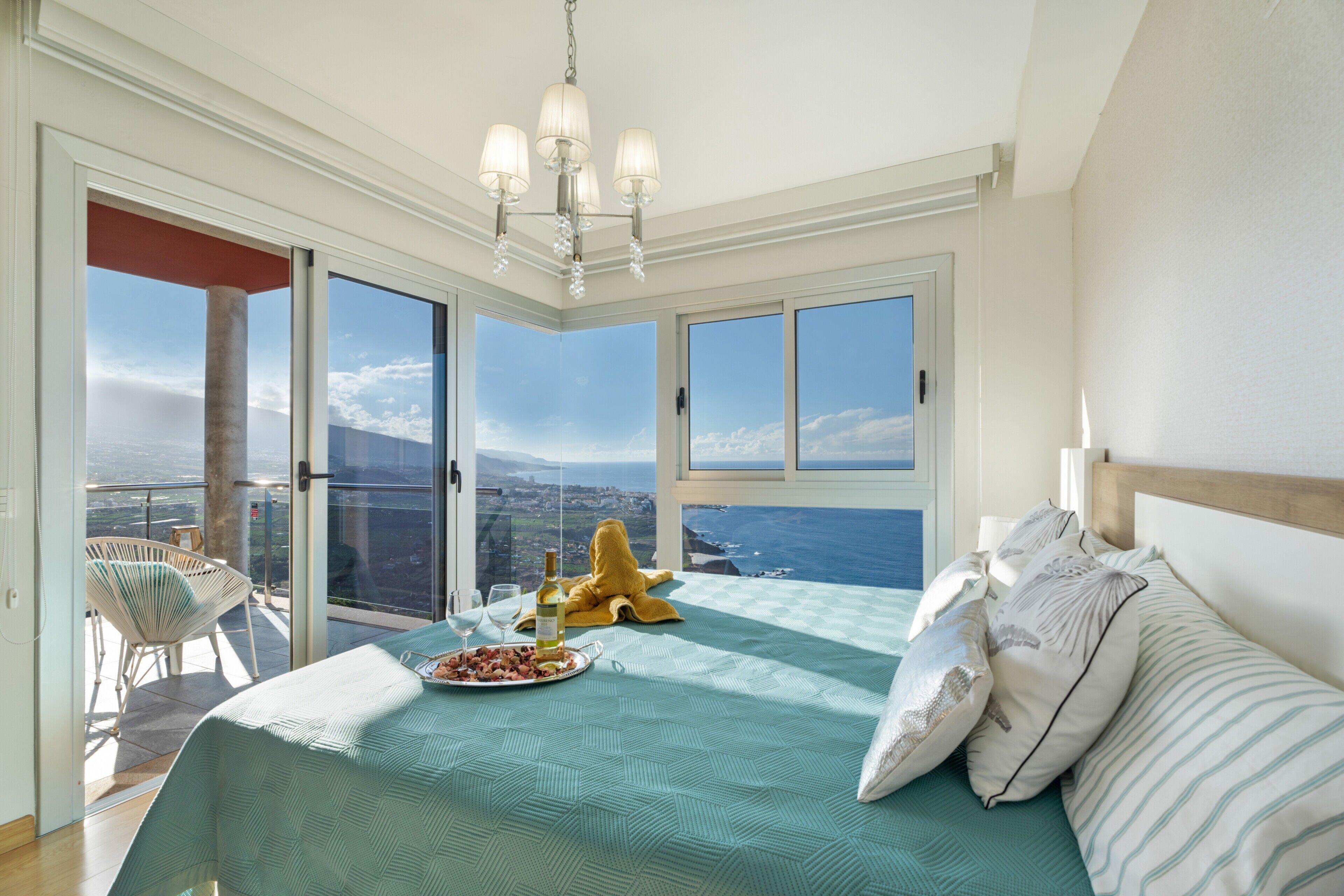 Une chambre à coucher avec un lit, un lustre, un balcon avec une chaise et une vue sur l’océan.