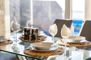 Una mesa de comedor con cubierta de vidrio transparente, platos blancos y cubiertos, adornada con individuales tejidos y jarrones decorativos.