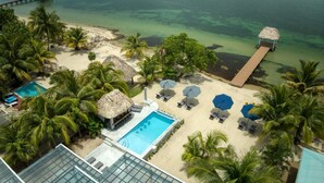 Vue aérienne d’un complexe hôtelier doté d’une piscine, de chaises longues, de parasols et d’une plage bordée de palmiers.