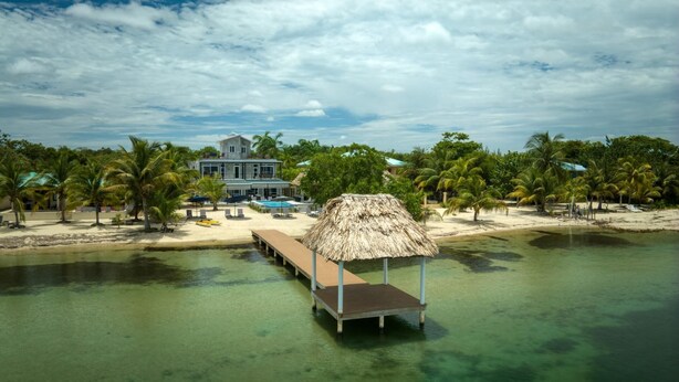 Une propriété située en bord de mer, dotée d’une structure à toit de chaume, d’une piscine et d’une grande maison avec de nombreuses fenêtres.